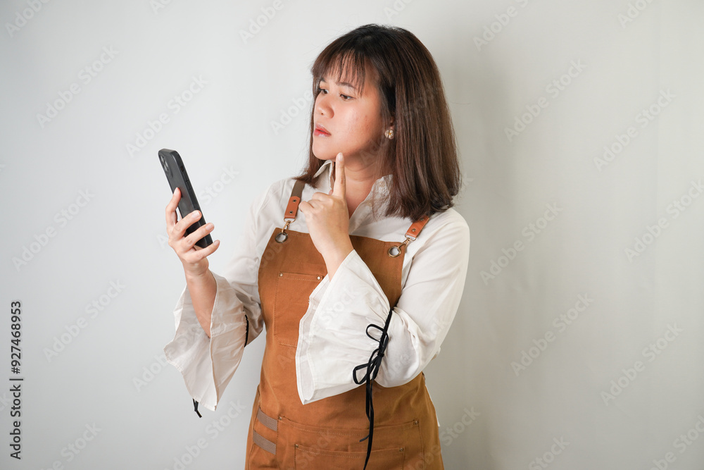 Curious young asian woman wearing long white sleeve shirt and brown apron is watching her smartphone with suspicious face expression. Isolated over white background. Curious young asian woman wearing long white sleeve shirt and brown apron is watching her smartphone with suspicious face expression. Isolated over white background.
