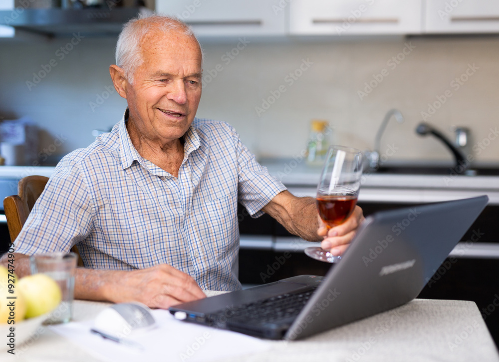 Elderly man with glass of wine communicates virtually using a laptop and the Internet Elderly man with glass of wine communicates virtually using a laptop and the Internet