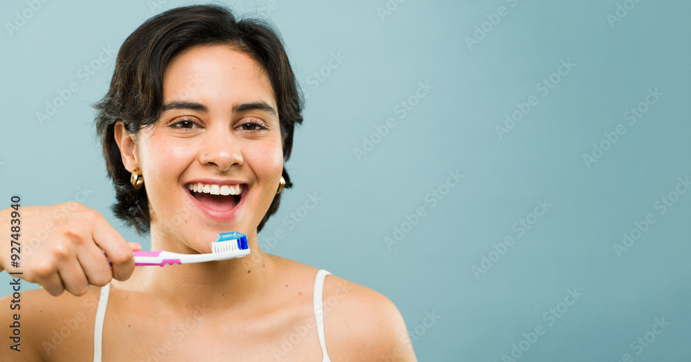 Happy Hispanic woman is happily brushing her teeth with a pink toothbrush on a blue background and plenty of copy space Happy Hispanic woman is happily brushing her teeth with a pink toothbrush on a blue background and plenty of copy space