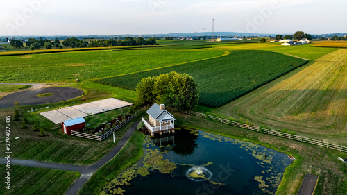 A tranquil rural landscape features a charming farmstead by a pond, surrounded by expansive green fields at sunset.