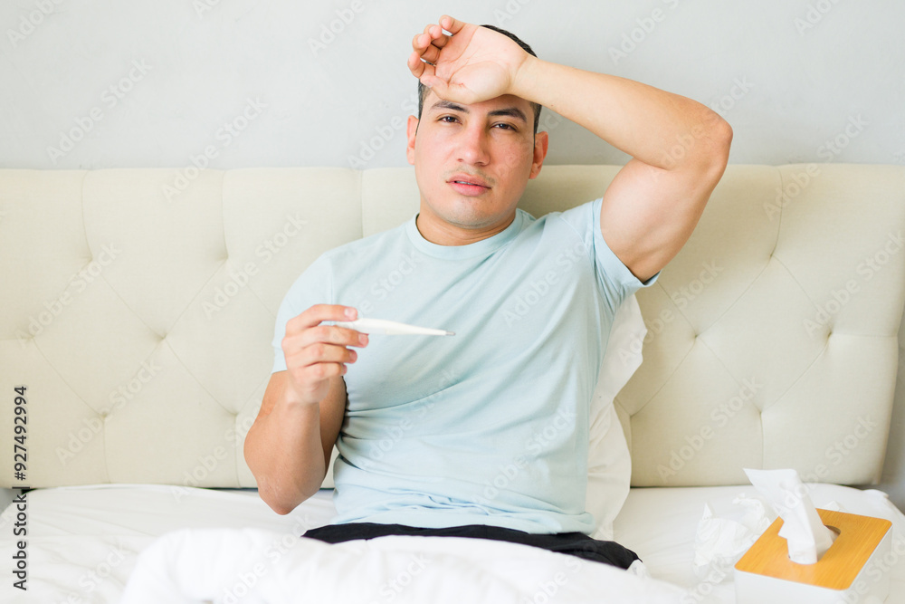 Man in bed feeling ill, checking his temperature with a thermometer Man in bed feeling ill, checking his temperature with a thermometer