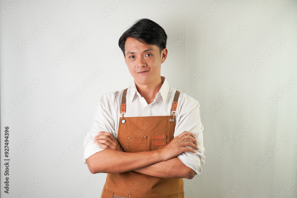 Confident young asian man wearing rolled-up white sleeve shirt and brown apron is smiling face expression to camera with arms crossed hand gesture. Isolated over white background. Confident young asian man wearing rolled-up white sleeve shirt and brown apron is smiling face expression to camera with arms crossed hand gesture. Isolated over white background.