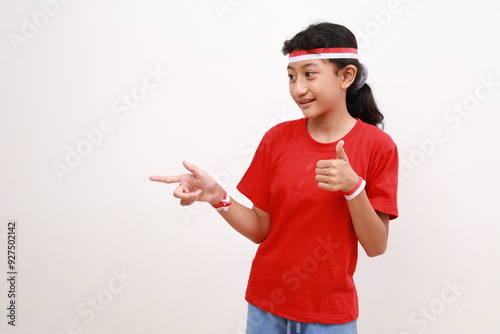 Happy young Asian girl in red white flag ribbon pointing fingers at copy space, showing thumb up gesture isolated on white background. Indonesian independence day on 17 august concept photo