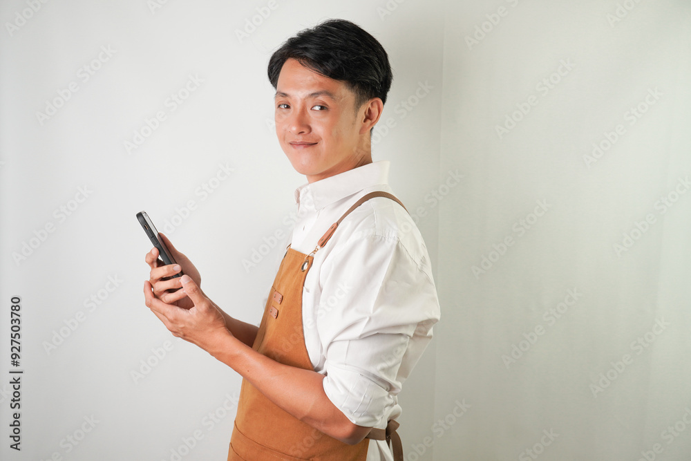 Confident young asian man wearing rolled-up white sleeve shirt and brown apron is holding a smartphone with smiling face expression. Isolated over white background. Confident young asian man wearing rolled-up white sleeve shirt and brown apron is holding a smartphone with smiling face expression. Isolated over white background.