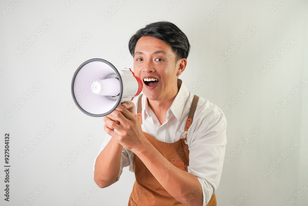Excited young asian man wearing rolled-up white sleeve shirt and brown apron is holding using megaphone with smiling face expression. Screaming, shouting at loudspeaker. Isolated over white background Excited young asian man wearing rolled-up white sleeve shirt and brown apron is holding using megaphone with smiling face expression. Screaming, shouting at loudspeaker. Isolated over white background