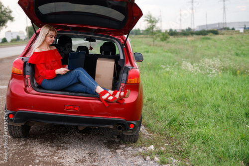 Young woman using laptop computer sitting in car trunk relaxing and enjoying her trip. Working while on vacation by car. Technology, freelancer, education online.