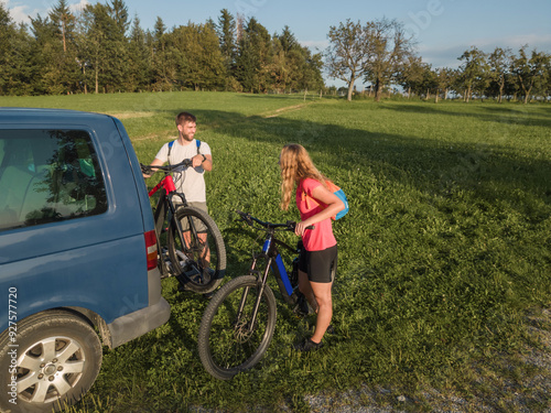 Young man and a woman preparing for off-road biking, taking down electric mountain bikes from the bike rack on the camper van. photo