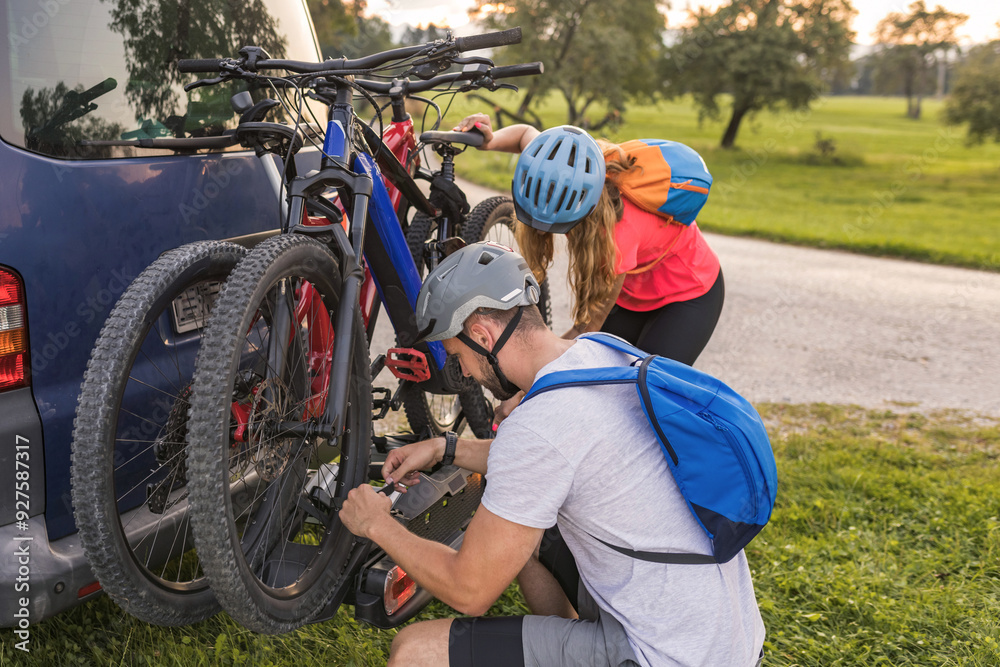 Fototapeta premium Young man is unloading electric mountain bikes, for himself and his girlfriend, lifting them from the hitch rack on the vehicle.