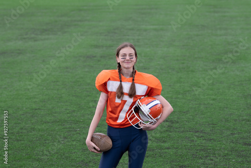 A smiling girl or young woman in an American football uniform is holding a ball and a helmet in her hands against the background of green grass on the football field.
