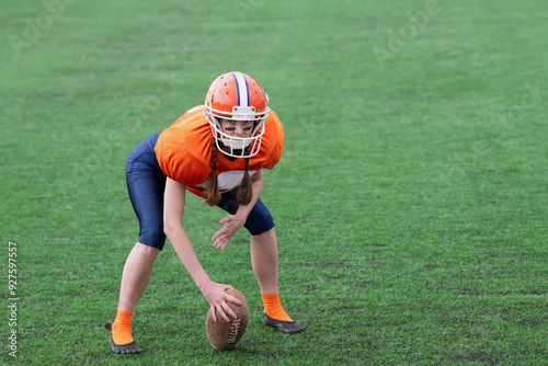 A girl or young woman in a helmet and an American football uniform is standing in the center position and holding the ball against the background of green grass on the football field.