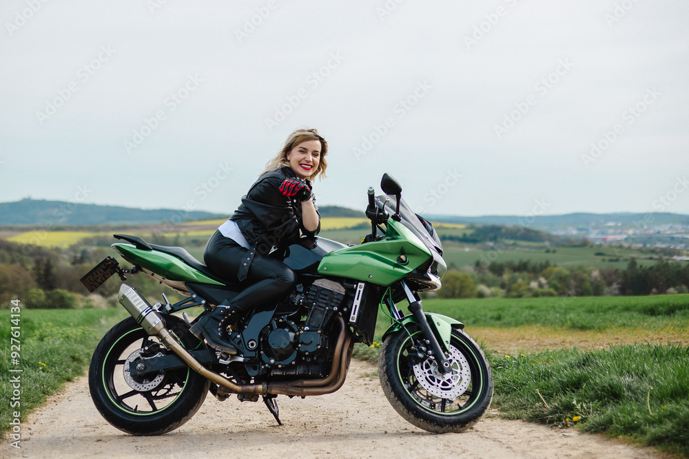 A woman in a leather jacket poses on a green motorcycle parked on a dirt road. The scene conveys a sense of adventure and freedom, with the rugged road and stylish bike. A woman in a leather jacket poses on a green motorcycle parked on a dirt road. The scene conveys a sense of adventure and freedom, with the rugged road and stylish bike.