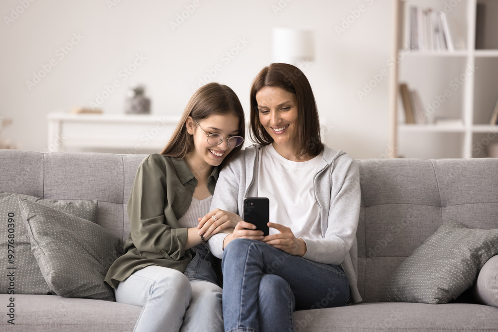 Smiling teenage girl spend time with mother sit on cozy sofa, have fun with mobile phone, use new application, watch video on social networks, read interesting news on internet, search for information Smiling teenage girl spend time with mother sit on cozy sofa, have fun with mobile phone, use new application, watch video on social networks, read interesting news on internet, search for information