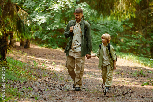 Father and son holding hands while walking through a lush forest, wearing casual hiking gear and carrying backpacks. Captured moment of bonding and adventure along a shaded trail