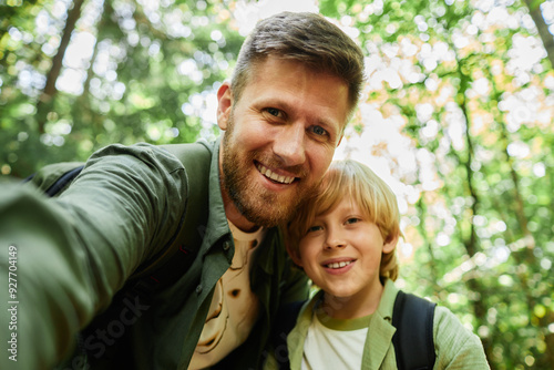 Portrait of father and son smiling for camera while taking selfie in lush green forest showing cheerful moment during outdoor adventure bonding experience photo