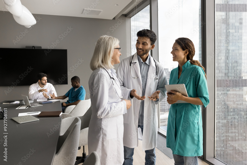 Three doctors, Indian, European and Hispanic internists in medical attire standing in modern clinic office, share opinion, skills, insights. Talk between experienced and young healthcare professionals Three doctors, Indian, European and Hispanic internists in medical attire standing in modern clinic office, share opinion, skills, insights. Talk between experienced and young healthcare professionals