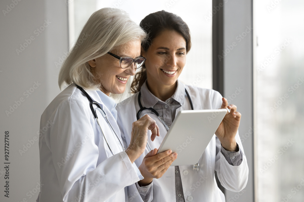 Two mature and young smiling female doctors reviewing new app or information on digital tablet, younger GP sharing details with older colleague. Mentorship, teamwork, consultation, knowledge-sharing Two mature and young smiling female doctors reviewing new app or information on digital tablet, younger GP sharing details with older colleague. Mentorship, teamwork, consultation, knowledge-sharing
