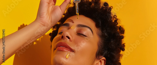 Beautiful black woman applying drops of oil serum on face using eye dropper, yellow background photo