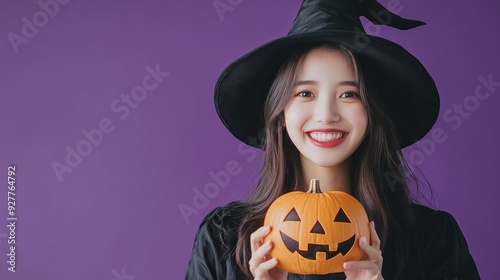 Halloween theme with a young Asian woman in a black costume wearing a witch hat smiling and holding a pumpkin lantern against a purple background