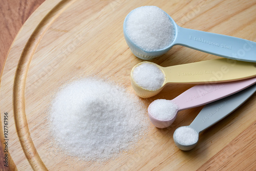 White granulated sugar in measuring spoon and pile of white granulated sugar on wooden plate for sugar level control and calorie control concept, glycemic control.
