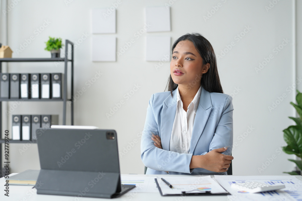 Businesswoman is sitting at her desk with her arms crossed, looking thoughtful while working on a project Businesswoman is sitting at her desk with her arms crossed, looking thoughtful while working on a project