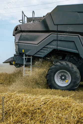 Obraz na płótnie Side view of a combine harvester in a wheat field, focusing on the machinery's tire and chaff dispersion. Advanced farming technology during harvest season. Agricultural efficiency. Vertical photo w salonie