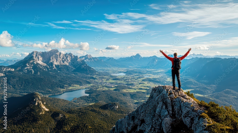 Obraz premium A breathtaking view from the mountain top, overlooking vast valleys, forests, and lakes. A single tourist stands at the peak with arms raised in triumph, wearing trekking gear and a backpack.