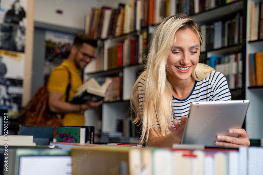 A cheerful woman engages with a tablet, while a man peruses bookshelves in a well-stocked library, both students immersed in their academic pursuits. A cheerful woman engages with a tablet, while a man peruses bookshelves in a well-stocked library, both students immersed in their academic pursuits.