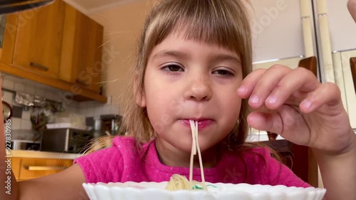 A European little girl in a pink T-shirt is eating long noodles in the kitchen. Close-up. High quality 4k footage