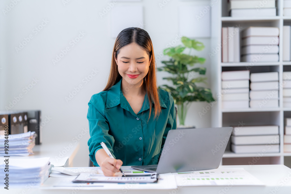 Focused and Efficient: A young Asian businesswoman, dressed in a vibrant green blouse, sits at her desk, immersed in work on a laptop and tablet, showcasing professionalism and dedication. Focused and Efficient: A young Asian businesswoman, dressed in a vibrant green blouse, sits at her desk, immersed in work on a laptop and tablet, showcasing professionalism and dedication.