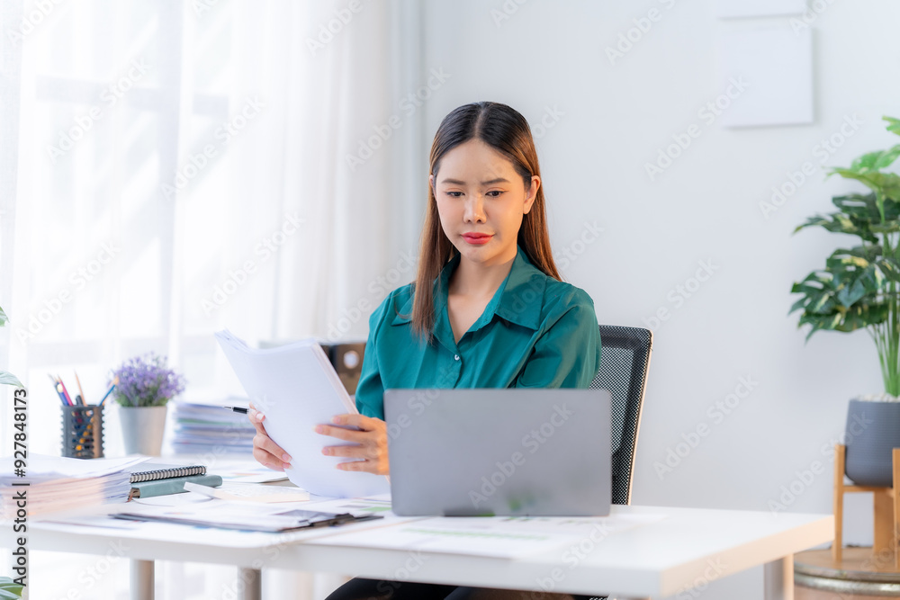 Focused Determination: A young woman in a green blouse sits at her desk, intently reviewing documents while working on her laptop. The soft, natural light and minimalist workspace create an atmosphere Focused Determination: A young woman in a green blouse sits at her desk, intently reviewing documents while working on her laptop. The soft, natural light and minimalist workspace create an atmosphere