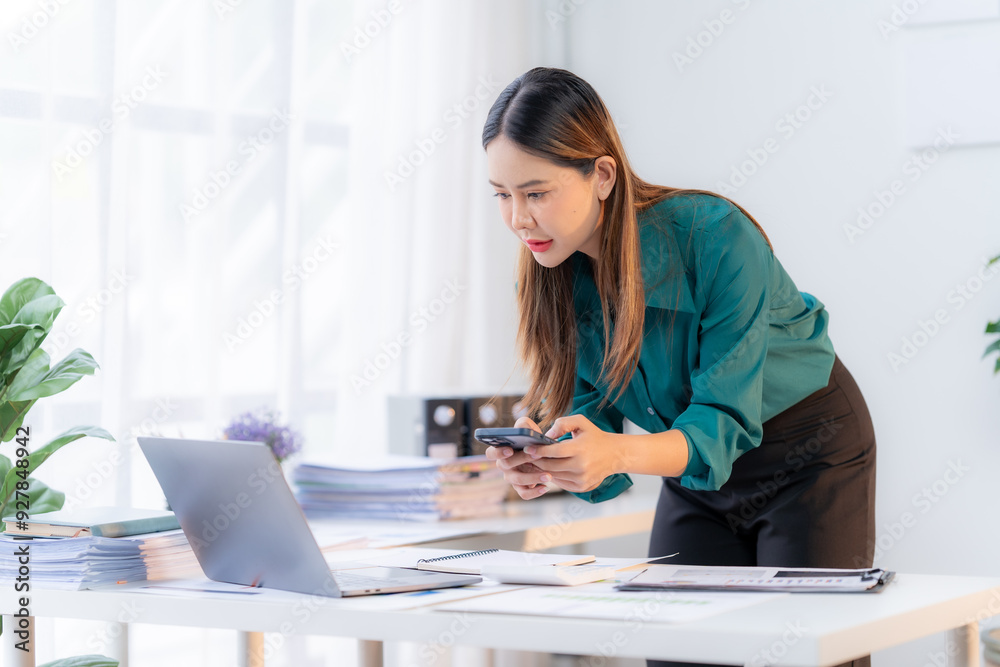 Focused Asian Businesswoman Analyzing Data: A young professional in a green blouse intently examines her phone while working on a laptop, embodying modern efficiency and productivity. Focused Asian Businesswoman Analyzing Data: A young professional in a green blouse intently examines her phone while working on a laptop, embodying modern efficiency and productivity.