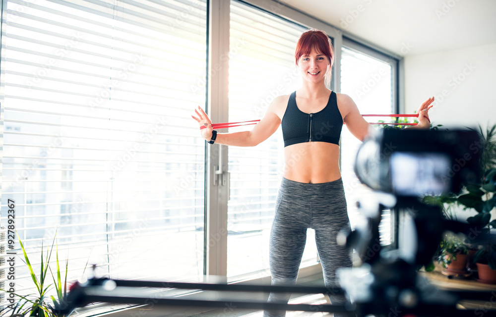 Fototapeta premium Redhead woman's home fitness routine with resistance band. Home workout on gym mat in living room, sporty woman wearing sportswear.