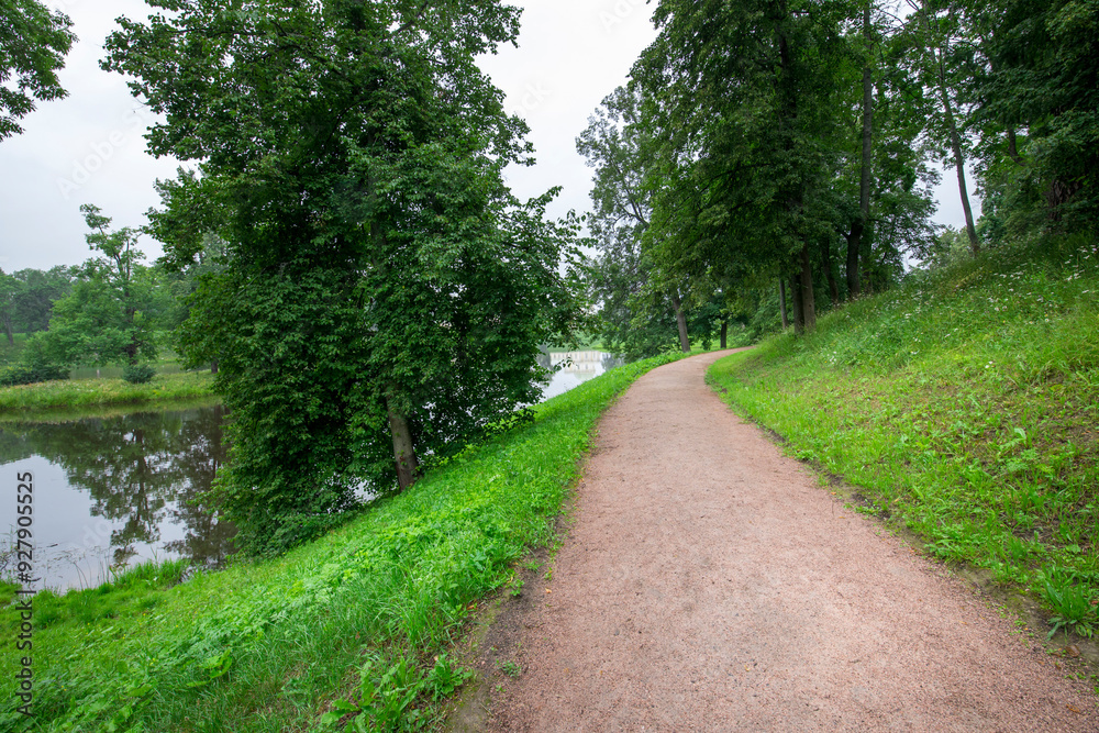A peaceful walkway meanders beside a calm lake, framed by vibrant green trees and soft grass, inviting a stroll in the refreshing morning atmosphere. A peaceful walkway meanders beside a calm lake, framed by vibrant green trees and soft grass, inviting a stroll in the refreshing morning atmosphere.