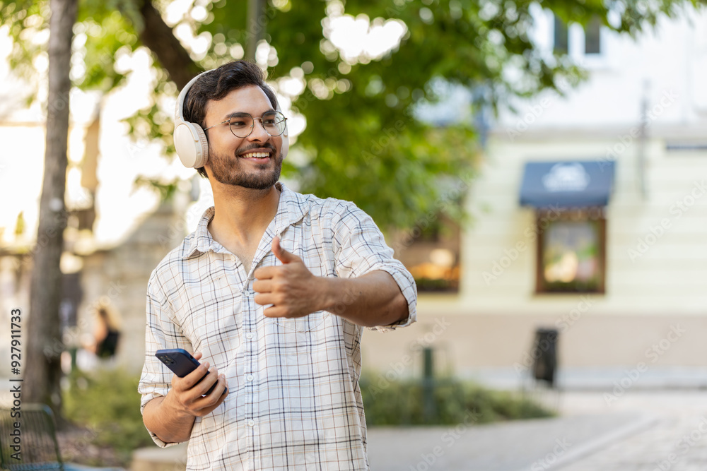 Happy overjoyed young man tourist in wireless headphones choosing, listening favorite energetic disco music on smartphone outdoors. Indian tourist guy standing on urban sunny city town street park. Happy overjoyed young man tourist in wireless headphones choosing, listening favorite energetic disco music on smartphone outdoors. Indian tourist guy standing on urban sunny city town street park.