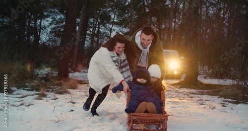 Happy European family has fun lying in snow on background of car in evening winter near pine forest. headlights turned on and warm glowing. Father mother with children playing together Christmas 4k