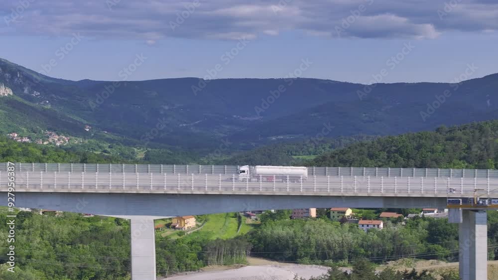AERIAL: White cargo truck drives along the spectacular Crni Kal viaduct on a sunny summer day. Scenic drone view of highway bridge running past beautiful karst mountain with a quiet hillside village. AERIAL: White cargo truck drives along the spectacular Crni Kal viaduct on a sunny summer day. Scenic drone view of highway bridge running past beautiful karst mountain with a quiet hillside village.