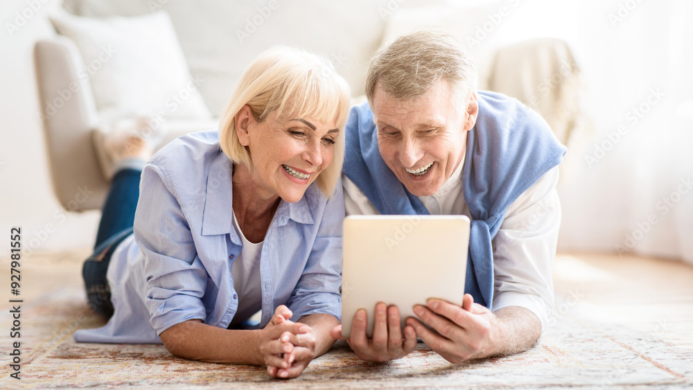 Excited mature couple browsing social media on tablet, lying on carpet at home Excited mature couple browsing social media on tablet, lying on carpet at home