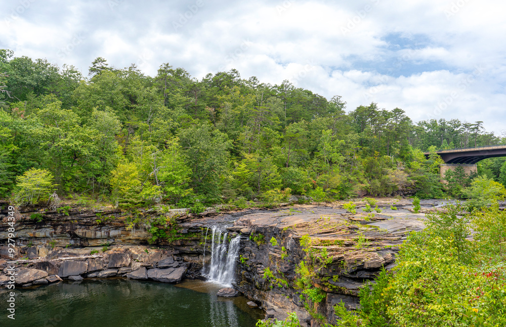 In the summer, Little River Falls in Little River Canyon National Preserve in Alabama is surrounded by green forests set against a cloudy blue sky. The Hwy 35 bridge is visible in the background. In the summer, Little River Falls in Little River Canyon National Preserve in Alabama is surrounded by green forests set against a cloudy blue sky. The Hwy 35 bridge is visible in the background.