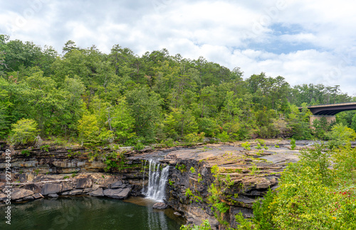 In the summer, Little River Falls in Little River Canyon National Preserve in Alabama is surrounded by green forests set against a cloudy blue sky. The Hwy 35 bridge is visible in the background.