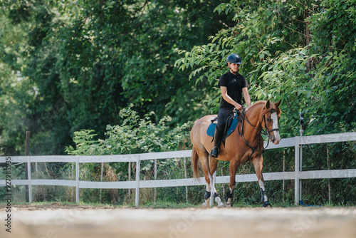 A woman wearing safety gear rides a horse along a fence-lined path in a lush, forested area on a bright sunny day.