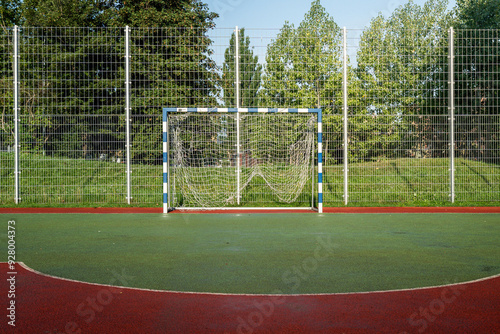 A soccer goal stands on a colorful outdoor pitch surrounded by a fence and greenery in broad daylight