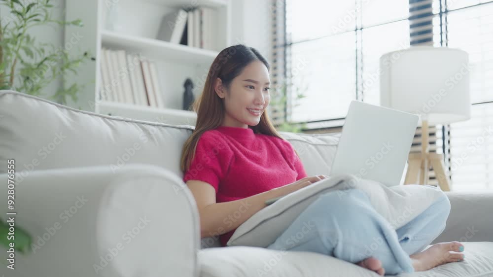 Happy young Asian woman using laptop while seated on couch at home Happy young Asian woman using laptop while seated on couch at home