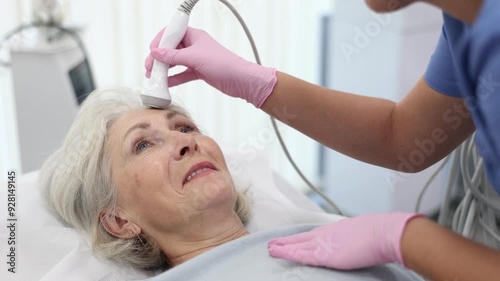 Relaxed aged woman having radiolifting of her face by means of apparatus for aesthetic procedures used by specialist  photo