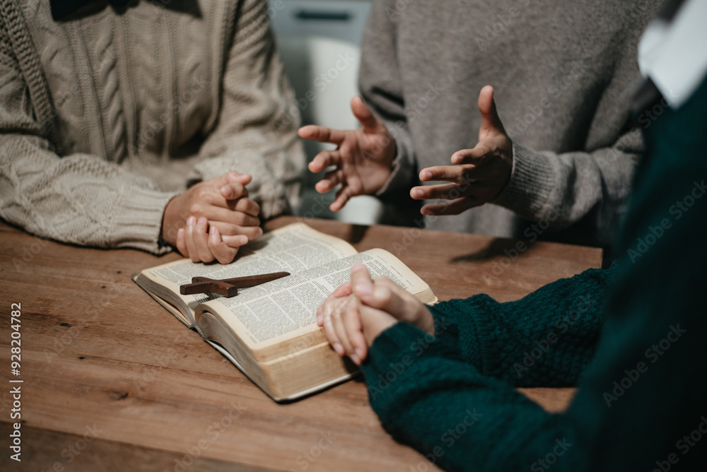 Group of Christians sit together and pray around a wooden table with blurred open Bible pages in their homeroom. Group of Christians sit together and pray around a wooden table with blurred open Bible pages in their homeroom.