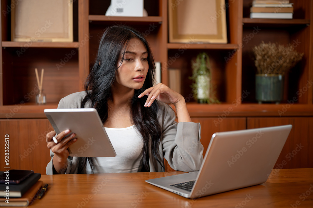 A businesswoman is focusing on her work, holding a tablet while reading online papers on laptop. A businesswoman is focusing on her work, holding a tablet while reading online papers on laptop.