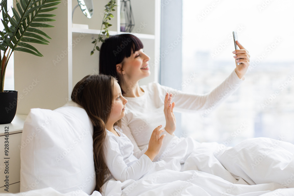 Morning routine with family connection and bonding time. Mom and daughter lying in bed having video call talking on phone.Bright, cheerful moment showing love and togetherness. Morning routine with family connection and bonding time. Mom and daughter lying in bed having video call talking on phone.Bright, cheerful moment showing love and togetherness.