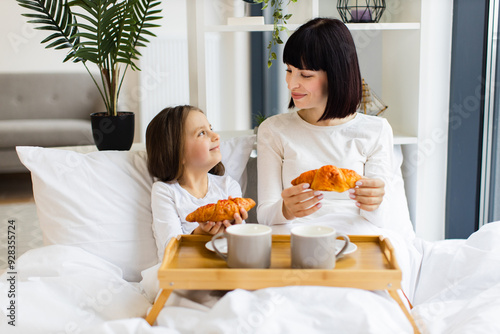 Mother and daughter sharing breakfast in bed, smiling at each other while holding croissants, bonding over a cozy morning. Wooden tray with croissants and cups on bed creates warm, intimate atmosphere photo