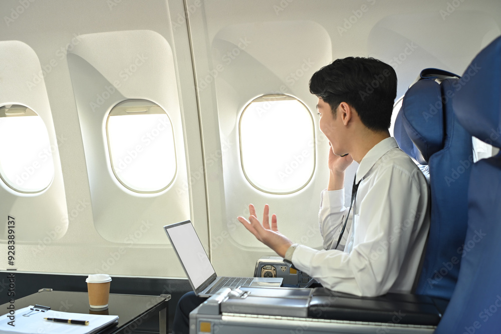 Fototapeta premium Side view of asian male passenger talking on his cellphone while sitting in an airplane seat with laptop