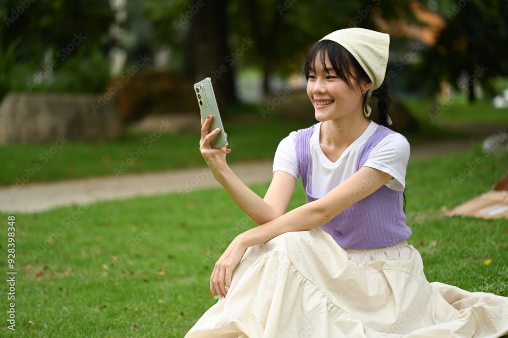 Fototapeta premium Pretty young asian woman sitting on the grass and using mobile phone