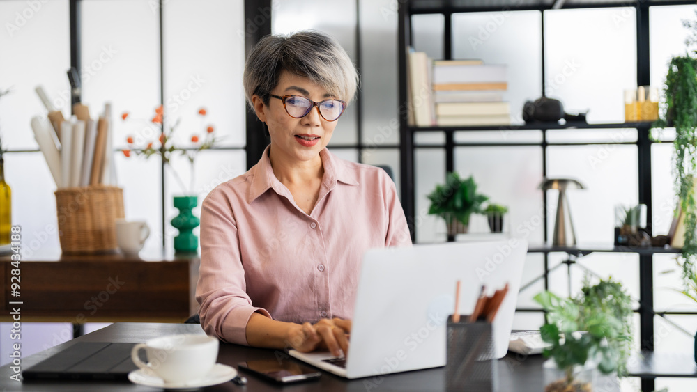 A middle-aged businesswoman with short gray hair is working on her laptop in a modern office, surrounded by a stylish, green-filled environment A middle-aged businesswoman with short gray hair is working on her laptop in a modern office, surrounded by a stylish, green-filled environment